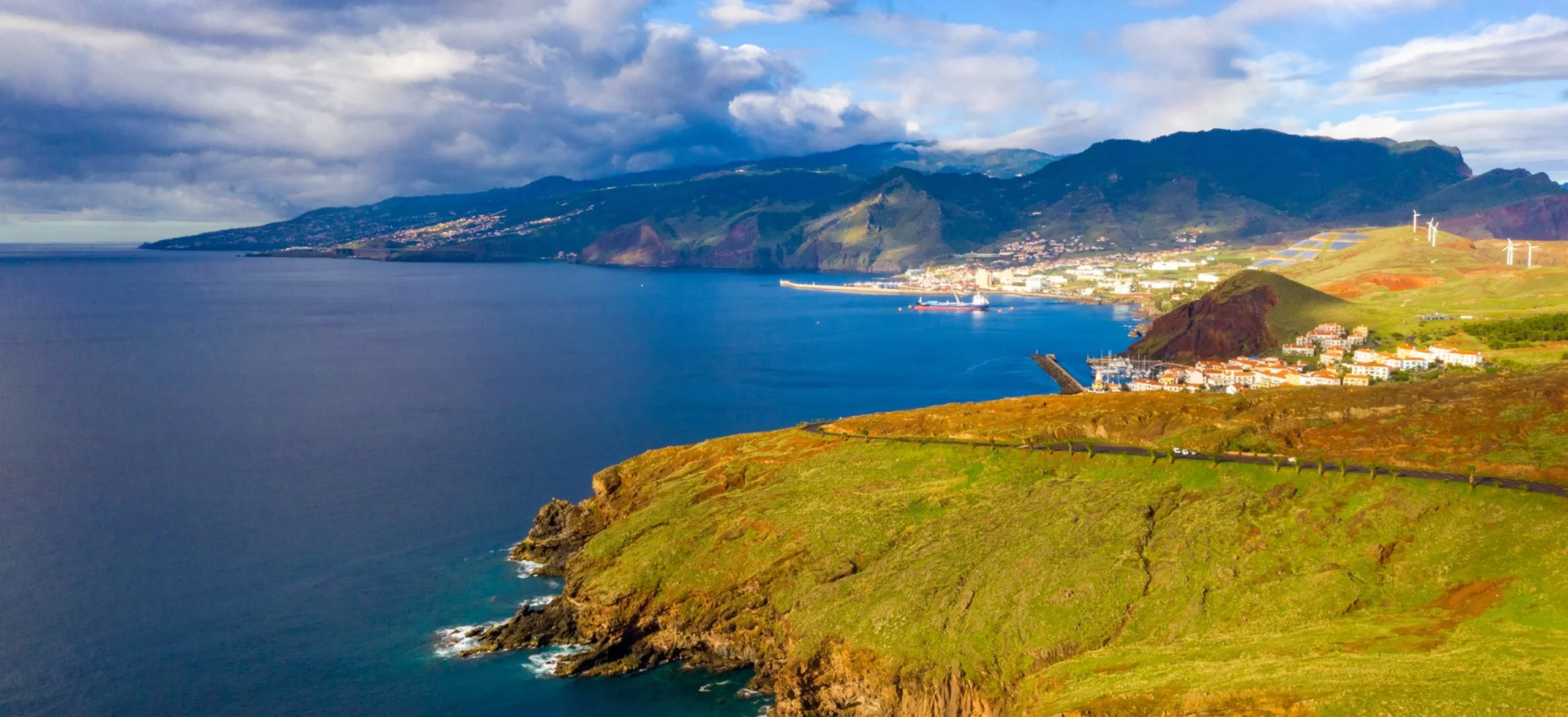 Panoramic aerial view of the rugged, eastern-most peninsula of Madeira (Ponta de São Lourenço) with wind turbines and a village in the distance.