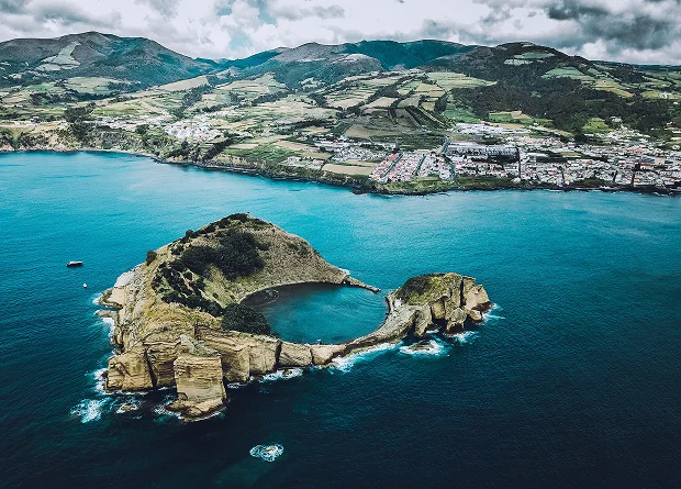Aerial shot of a unique, almost perfectly circular volcanic crater islet with a turquoise lagoon inside, located off a lush green island coastline.